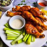 Buffalo chicken drumsticks arranged on a plate with a small dish of blue cheese and creamy salad in the background