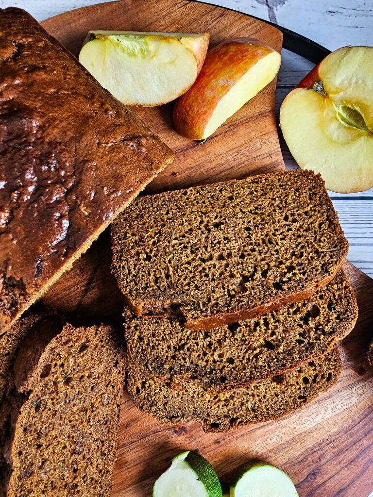 UP CLOSE SLICES OF ZUCCHINI APPLE BREAD SERVED ON A WOODEN CUTTING BOARD WITH SLICED APPLED ON THE SIDE