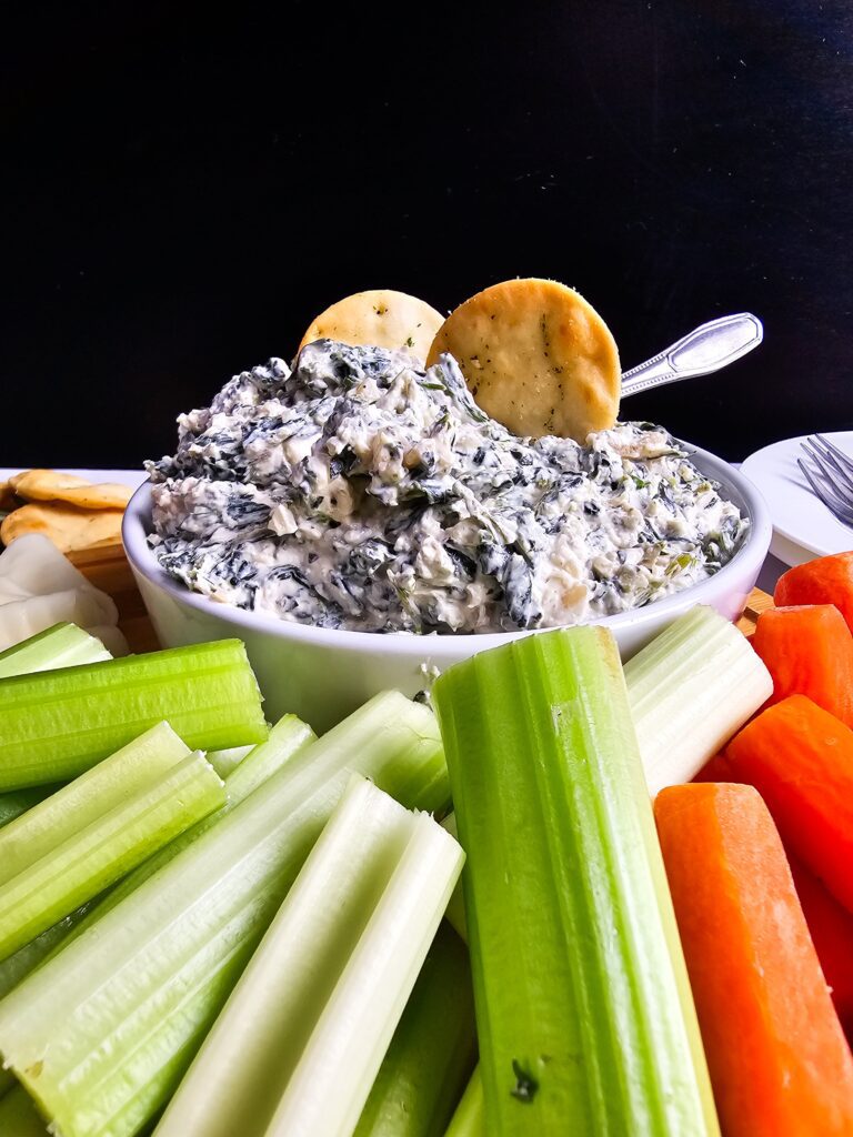 UP CLOSE BOWL OF SPINACH DIP SUROUNDED BY CARROTS, CELERY AND CRACKERS.