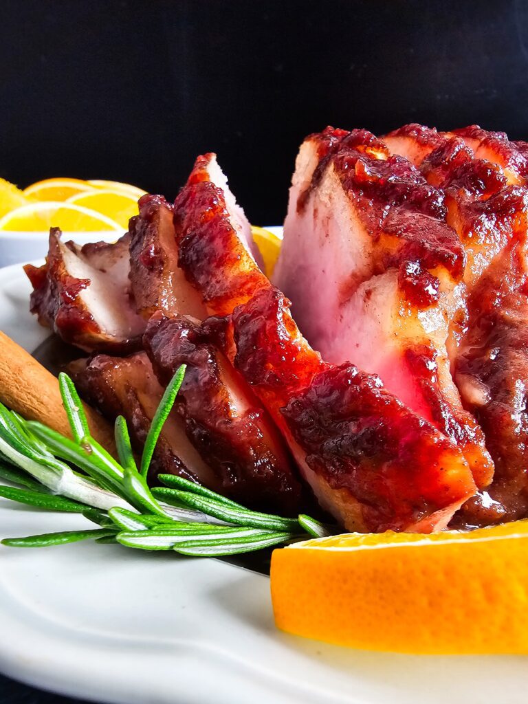 UPCLOSE PHOTO OF THE GLAZED HAM SLICED ON A FANCY SERVING PLATTER GARNISHED WITH ROSEMARY AND SLICED ORANGES