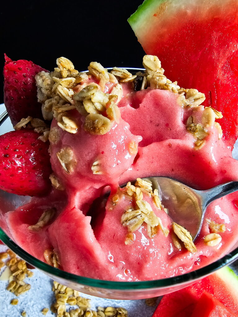 Close up of thick watermelon strawberry smoothie bowl showing spoonable ice cream like texture topped with crunchy granola and fresh strawberries with a spoon digging in
