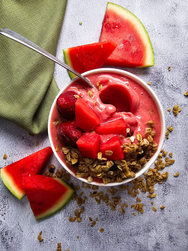 Watermelon smoothie bowl with fresh watermelon cubes strawberries and crunchy granola in a white bowl surrounded by watermelon slices on a grey surface
